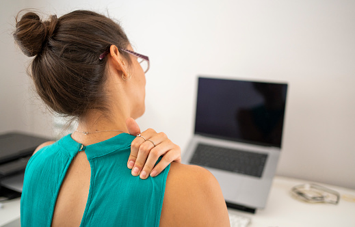 Rear view of a young businesswoman massaging her sore shoulder while working on a laptop at her office desk