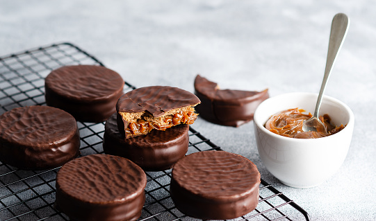 Dark chocolate alfajores on a dark rack and a bowl with dulce de leche, on grey background.
