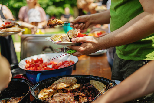 Group of colleagues serving food during team building picnic, promoting friendship outside the office. Lots of barbecue, tomatoes, bread, on a wooden table on the grass, in the shades of a tree, with unrecognizable people choosing what to eat.