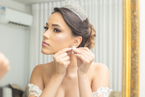 Goiania, Goiás, Brazil – May 15, 2022:  Young woman in a wedding dress, in front of the mirror, putting on her earrings.