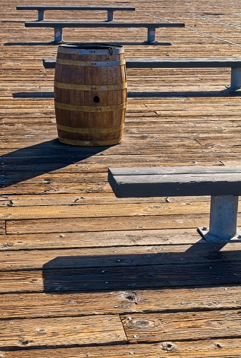 Park bench and trash barrel on wooden pier at Fisherman's Warf, San Francisco. Old wooden planks, benches and oak barrel interplay with shadows at this famous pier.