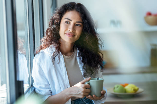 istock Beautiful woman drinking a cup of coffee while looking forward standing next to the window at home. 1418282639
