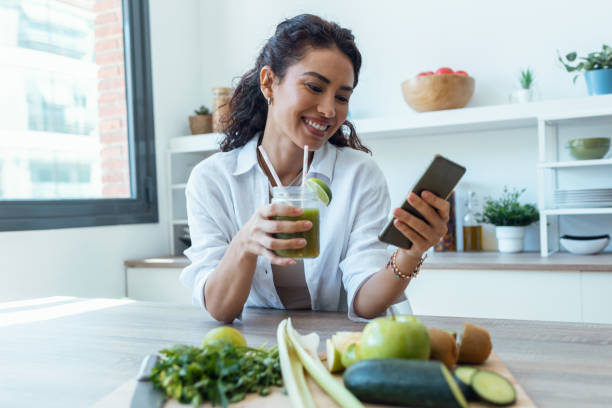 симпатичная женщина использует свой мобильный телефон во время питья фруктового детокс-сока на кухне дома. - smart casual occupation casual healthy eating стоковые фото и изображения