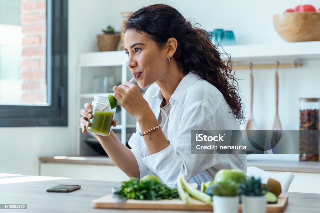 Pretty woman looking to sideways while drinking fruit detox juice in the kitchen at home. Shot of pretty woman looking to sideways while drinking fruit detox juice in the kitchen at home. Healthy Eating Stock Photo