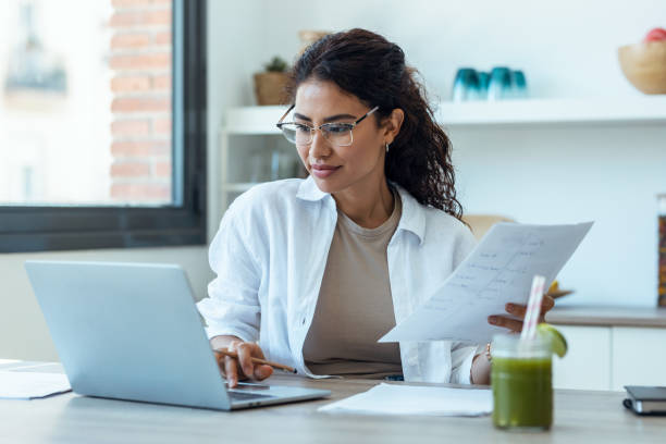istock Pretty business woman working with computer while consulting some invoices and documents in the kitchen at home. 1418283270