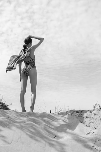 young woman in swimwear and cloak stand on sand dune under wind looking at horizon, rear view, monochrome