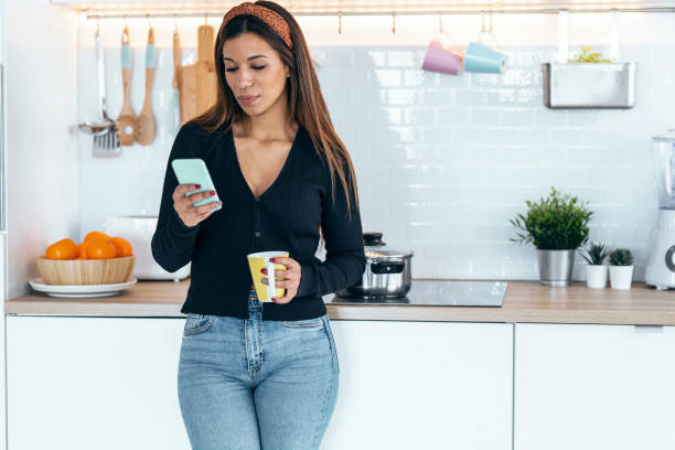 istock Beautiful woman using her mobile phone while drinking a cup of coffee in the kitchen at home. 1418289971