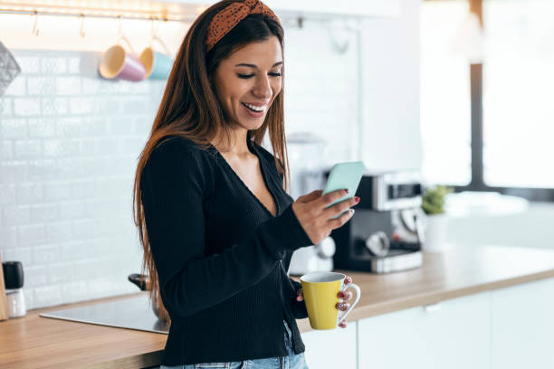 istock Beautiful woman using her mobile phone while drinking a cup of coffee in the kitchen at home. 1418290499