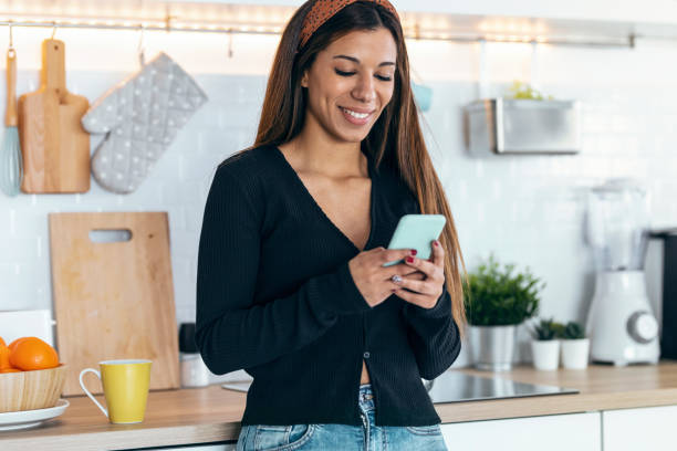 istock Beautiful woman using her mobile phone while standing in the kitchen at home. 1418290600