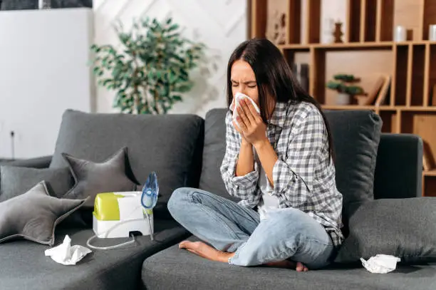 Sick caucasian brunette girl sit on couch at home, feel unhealthy, she suffering from rhinitis snuffles, having respiratory infection, get flue,  sitting at home at quarantine, need treatment