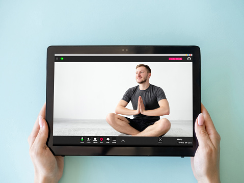 Yoga video class. Online meditation. Virtual training. Woman hands holding tablet with relaxed man in lotus pose on screen isolated on blue.