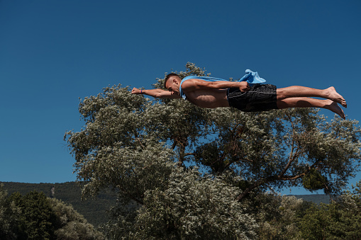 Young teen boy wearing a towel as a superhero scarf flying and diving in river. Clear blue sky and trees in distance as a natural background. High quality photo