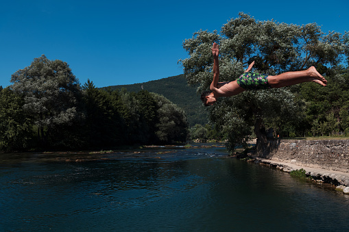 Young teen boy jumping flying and diving in the river. Clear blue sky and trees in distance as a natural background. High quality photo