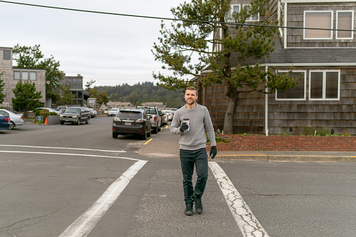 A mature, thirty-three year old man is walking across the street smiling with a coffee in hand. He's looking off camera and is wearing a grey sweater and fall gloves.