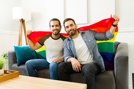 Portrait of a happy gay man and his partner supporting LGBT rights and waving a rainbow flag while at home