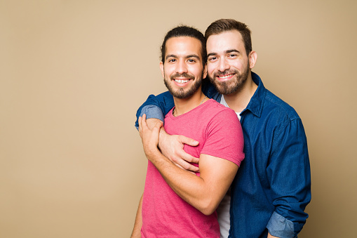 Attractive happy gay couple hugging and smiling while looking at the camera in front of a studio background with copy space