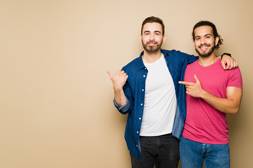 Smiling attractive gay couple in casual clothes pointing next to copy space ad in front of a studio background