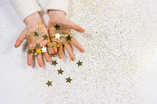 Child's hands holding golden stars with golden glitter on light backgound. Christmas magic and celebration. Festive atmosphere, holiday concept.