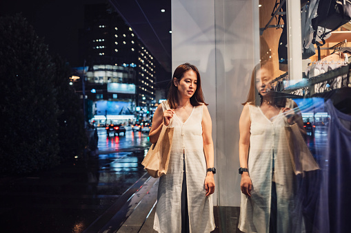 Asian woman standing outside a boutique looking at shop window in the evening in the city