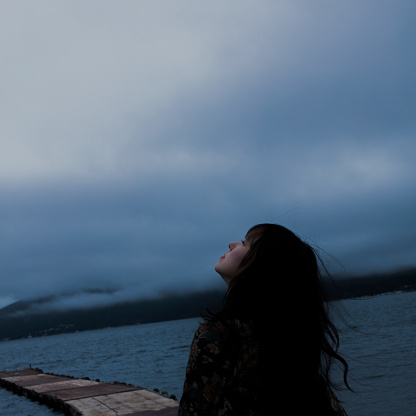 A square photo showing the upper body of a Japanese woman with long hair looking up at the sky by the lake on a cloudy day.