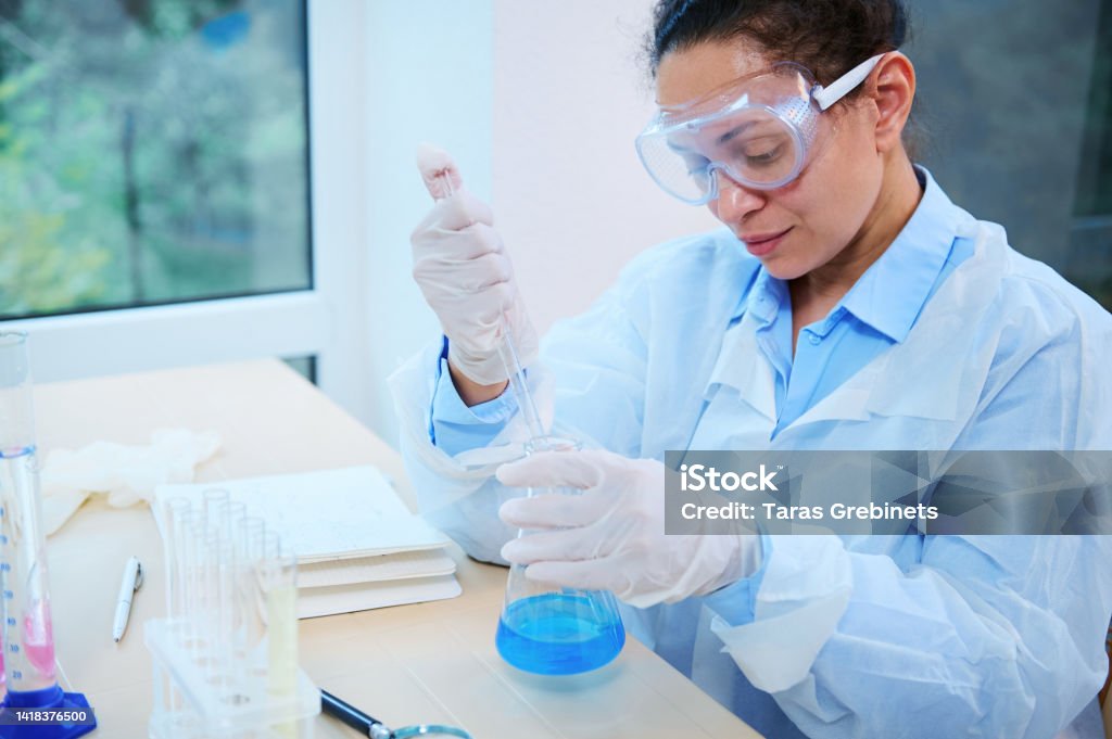 A woman scientist pharmacologist pipetting blue chemical liquid in flat-bottomed flask, conducting a science experiment African American woman, scientist pharmacologist pipetting the blue chemical liquid in a flat-bottomed flask, sitting at a table in a light laboratory interior, conducting a science experiment Adult Stock Photo