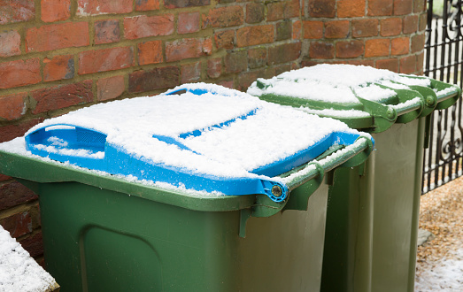 Green and blue wheelie bins in a garden in winter, UK. Bin lids covered in snow.