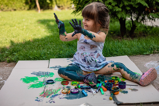 The child is outdoor, playing with some watercolors. She is happy, because she can use her fingers to create the art.