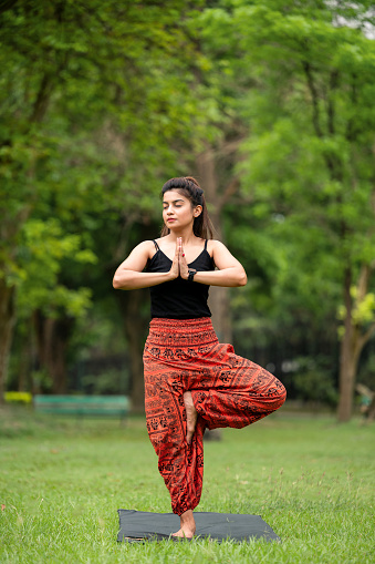 Outdoor image of young Athletic slim woman with hair bun in tight sportswear practicing yoga, doing Vrikshasana or Tree Pose exercise, standing on one leg in Namaste position.