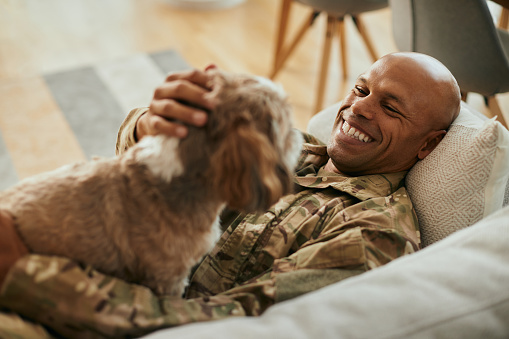 Young happy African American veteran cuddling his dog while relaxing on the sofa.