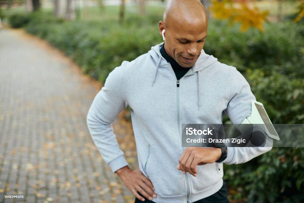 Young black athlete using fitness tracker while exercising in the park. Happy African American sportsman using smart watch while working out in nature on autumn day. Autumn Stock Photo