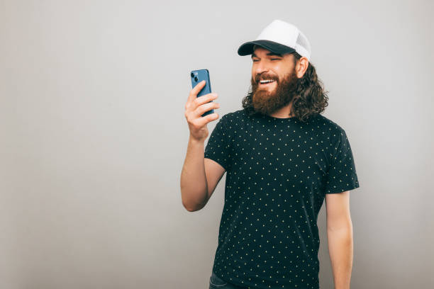 Happy bearded man is texting with someone smiling while he stands near a gray wall stock photo