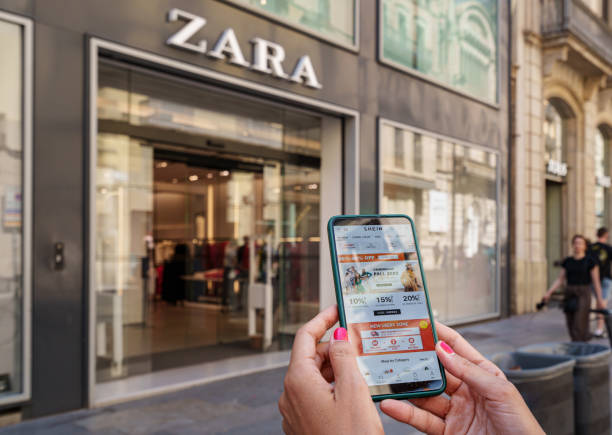Woman holding a smartphone with the Shein app on the screen and a Zara store in the background stock photo