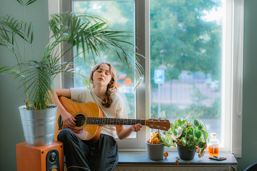 Young Caucasian woman sitting on windowsill and playing guitar