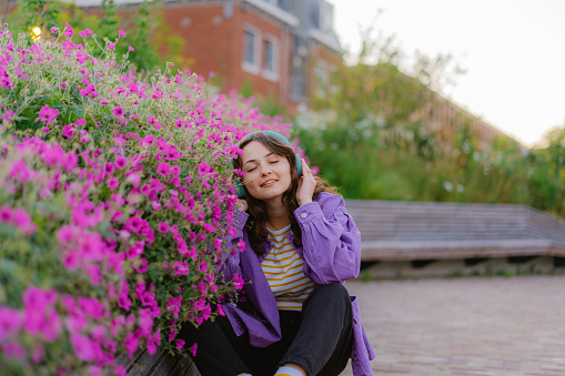 Young Caucasian woman in purple coat  listening to music in headphones on the background of purple flowers