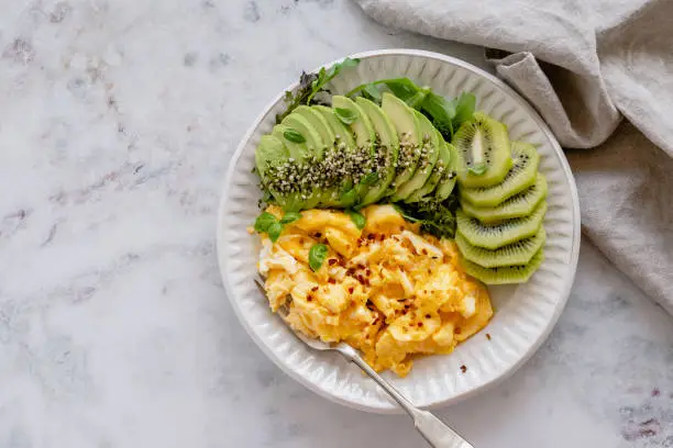Overhead view of a plate of scrambled eggs with a side of avocado, salad and sliced kiwi