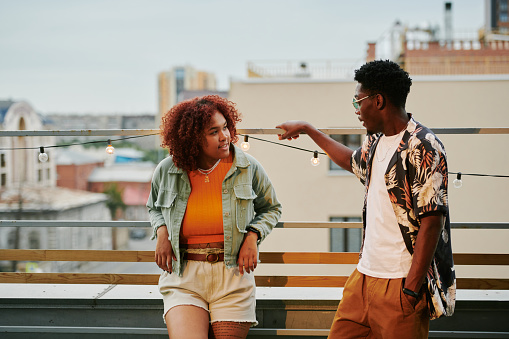 Young African American couple in stylish casualwear having chat on terrace of rooftop cafe against urban environment