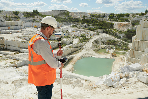 Side view of mature engineer or surveyor adjusting theodolite before measuring territory of marble quarry on summer day