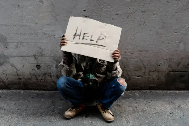 Photo of A homeless, long-haired Asian man sits hopelessly leaning against a wall as there is no one to help him with work and food in his hand holding a sign for help. Homeless sleep on streets