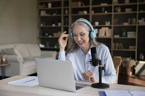 Happy senior blogger woman in big wireless headphones speaking at professional mic and laptop, giving workshop, smiling, laughing, broadcasting, recording podcast. Communication concept