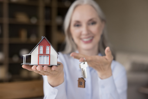 Happy mature mortgage broker woman holding keys and small model of house on hand. Real estate agent, real estate agent offering apartment for rent, selling, buying. Property purchase concept