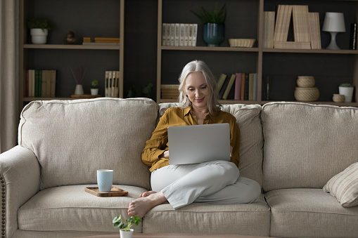 Happy engaged retired grey haired lady resting on couch at home, enjoying leisure, entertainment, using laptop on comfortable sofa, watching moviel typing, chatting, working