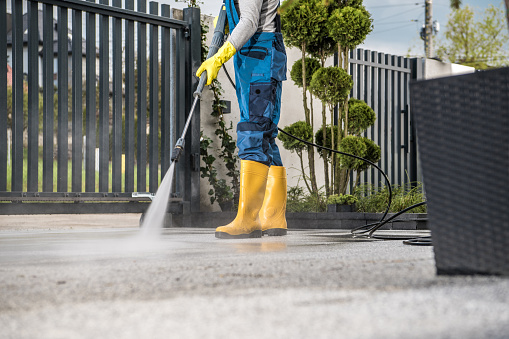 Man in Yellow Rain Boots and Work Uniform Pressure Washing Concrete Tiles of the Driveway to His House Behind the Closed Yard Entrance Gate. Home Surroundings Maintenance Theme.