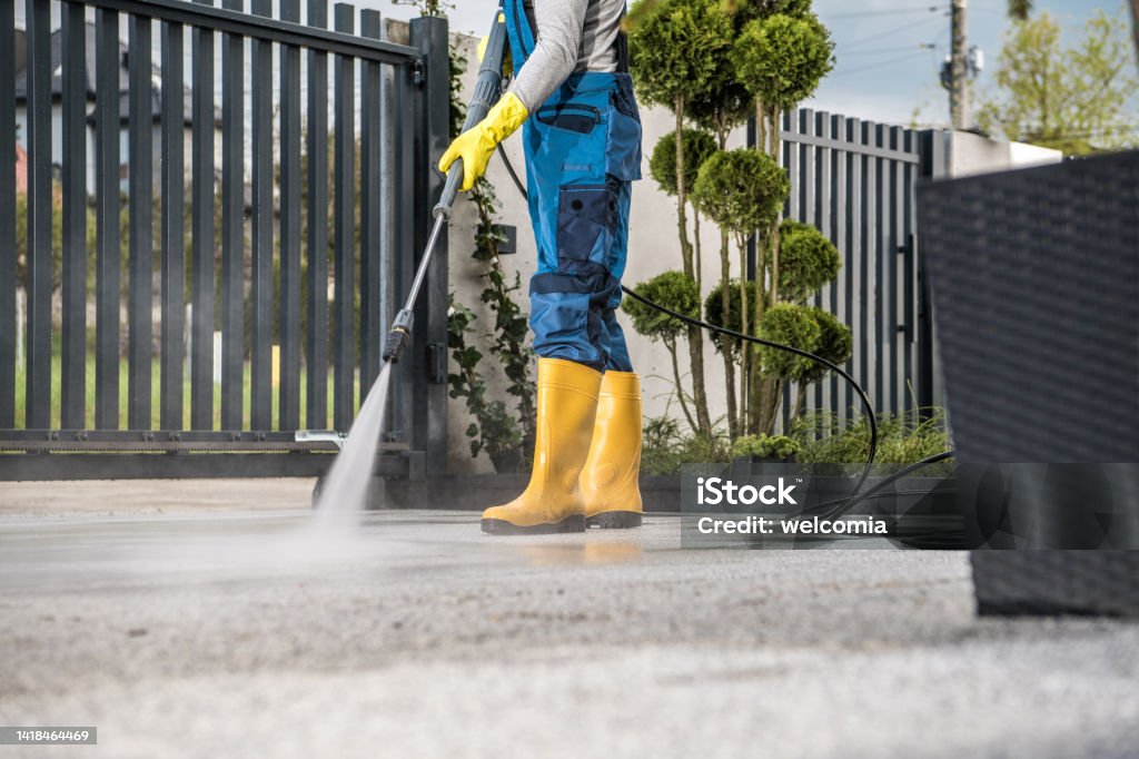 Cleaning the Driveway with Pressure Washer Man in Yellow Rain Boots and Work Uniform Pressure Washing Concrete Tiles of the Driveway to His House Behind the Closed Yard Entrance Gate. Home Surroundings Maintenance Theme. High Pressure Cleaning Stock Photo