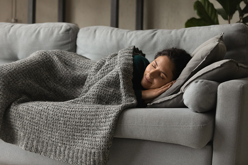 Happy mindful millennial generation hispanic caucasian woman napping daydreaming on comfortable sofa under warm blanket, sleeping on soft pillow alone in modern living room, peaceful weekend pastime.