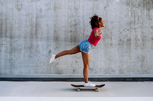 Multiracial teenage girl riding skateboard in front of concrete wall, standing in one leg, balancing. Side view.