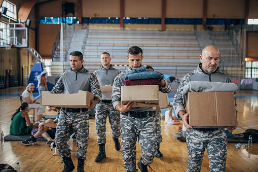 Group of people, soldiers on humanitarian aid carrying donation boxes for civilians in school gymnasium, after natural disaster happened in city.