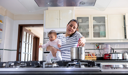 Latin American mother multi-tasking at home cooking and talking on the phone while carrying her baby - lifestyle concepts