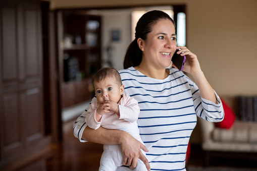 Happy Latin American mother multi-tasking at home talking on the phone while carrying her baby - lifestyle concepts