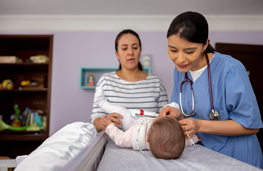 Latin American female doctor making a house call visit and checking on a baby girl wearing a hip dysplasia brace in front of her mother - pediatric medicine concepts