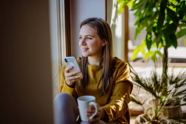 istock Young college student using phone, when sitting on floor at home and enjoying cup of tea. 1418488065